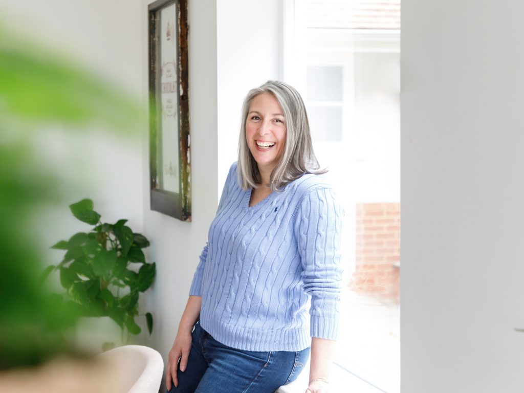Janet Padfield, Nutritional Therapist in a blue jumper and leaning on a window sill.